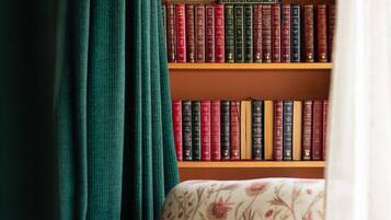 Una libreria piena di libri, una poltrona con motivo floreale e una tenda verde che copre parzialmente la vista.