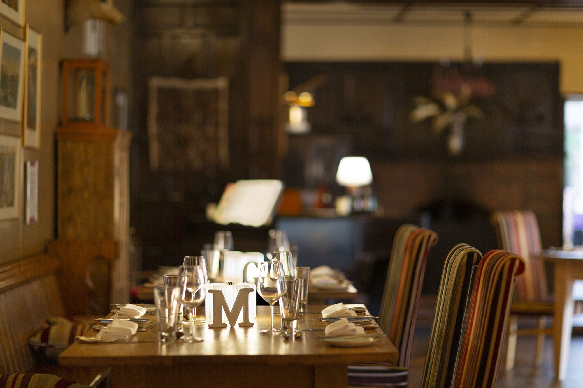 Mesa de comedor dispuesta con copas de vino, un centro de mesa con la letra 'M' y sillas a rayas.