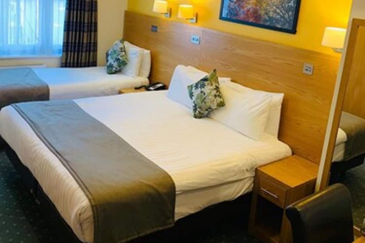 A hotel room with two beds, a wooden headboard, a small wooden nightstand, a table, and a window with curtains.