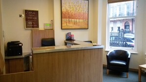 A reception area with a wooden reception desk, a black office chair, a painting on the wall, and a window with a view of the street. - Regency Hotel Parkside (London)