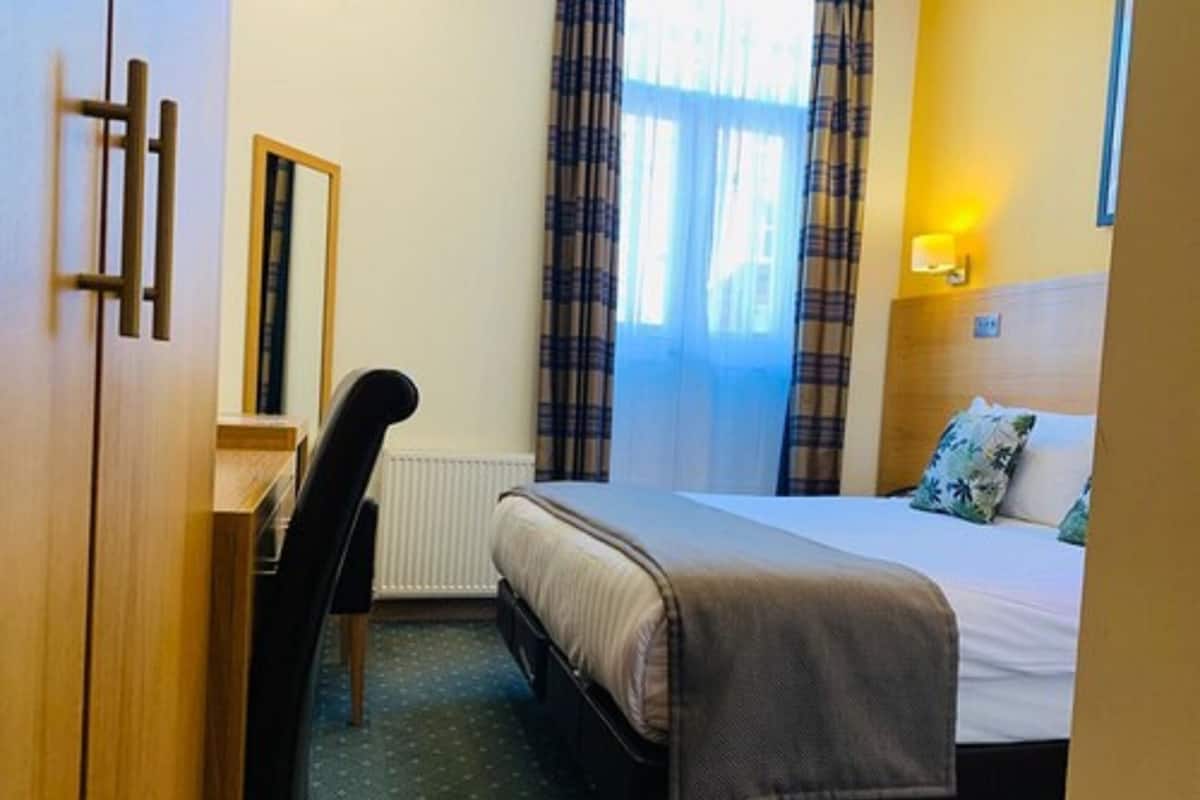 A hotel room with a bed, a desk, a chair, a window with curtains, and a wall-mounted light.