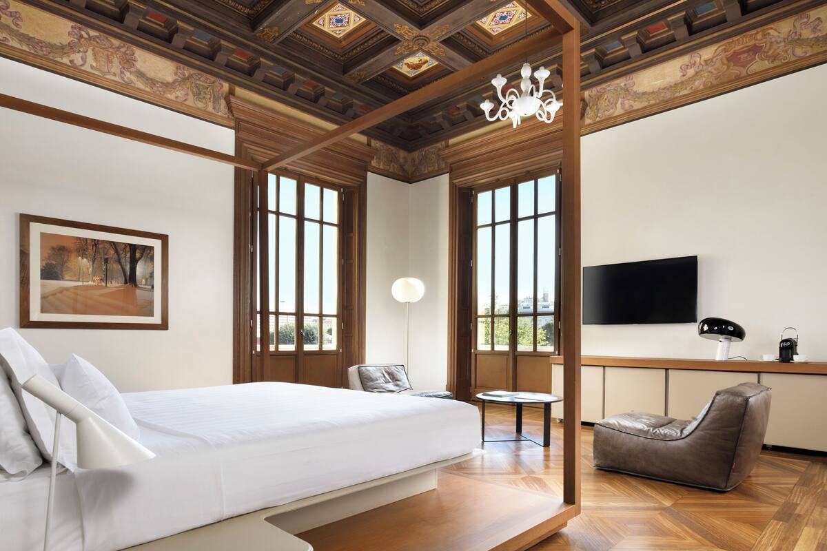 A modern hotel room with a large bed, a seating area, a television, and a decorative ceiling.