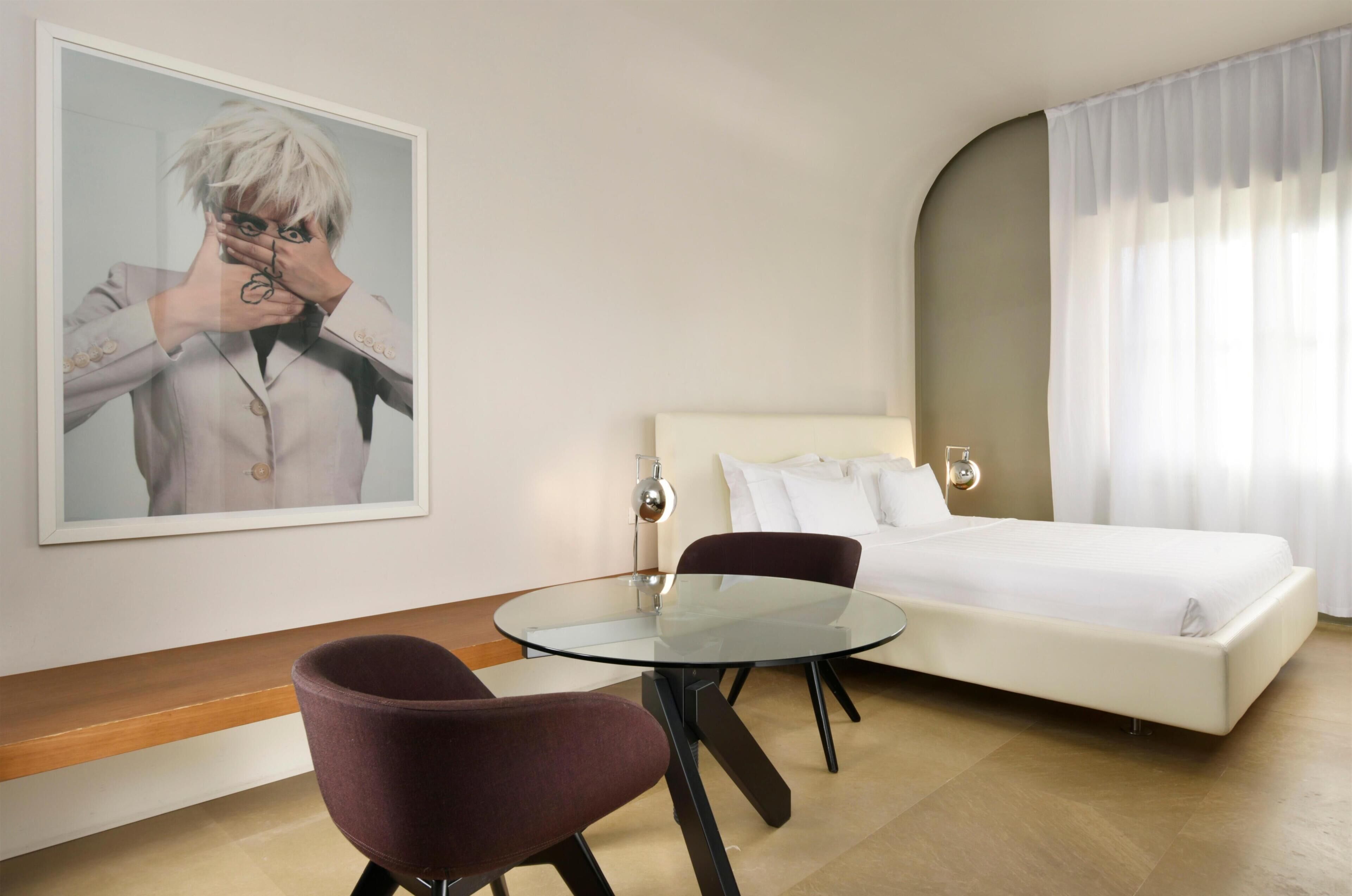 A hotel room with a bed, a round glass table, a chair, a painting on the wall, and a window with curtains.
