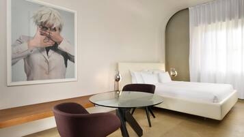 A hotel room with a bed, a round glass table, a chair, a painting on the wall, and a window with curtains.