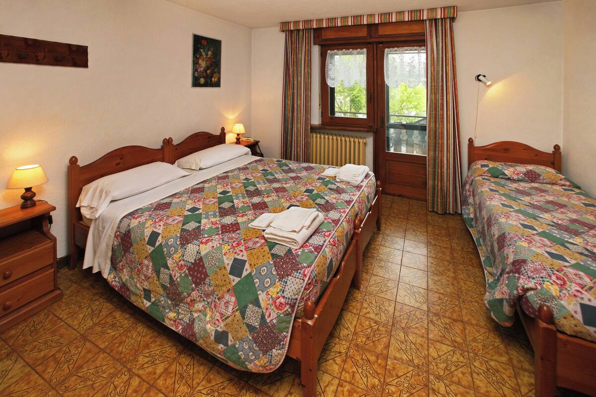 A bedroom with two beds, a wooden headboard, a quilt, a nightstand, and a window with curtains.
