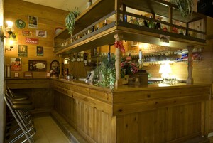 Angolo bar in legno con scaffali pieni di bottiglie, un bancone con bicchieri e una zona con sgabelli.