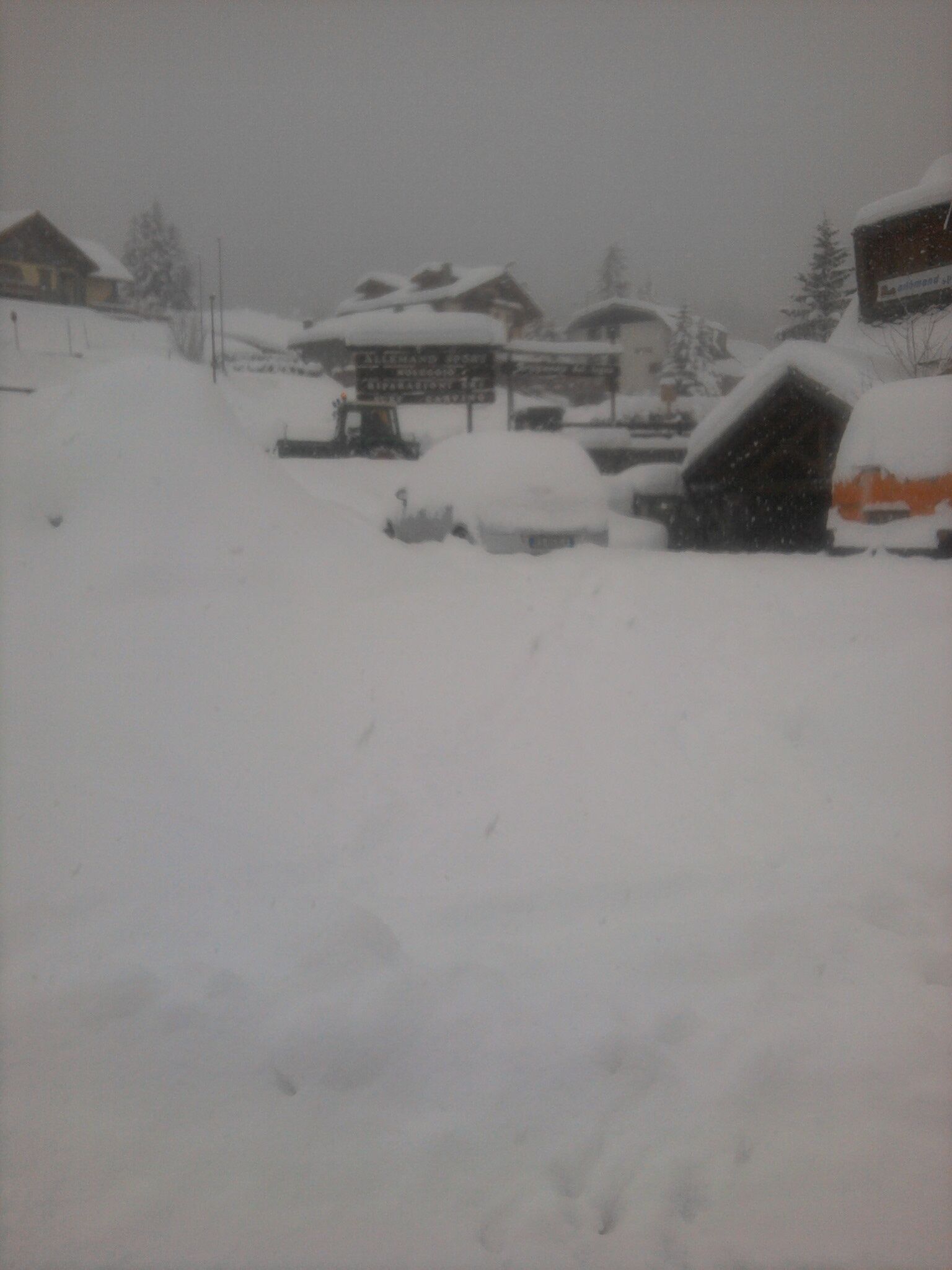 Escena nevada con un edificio cubierto de nieve, un cartel y un vehículo semienterrado en la nieve.
