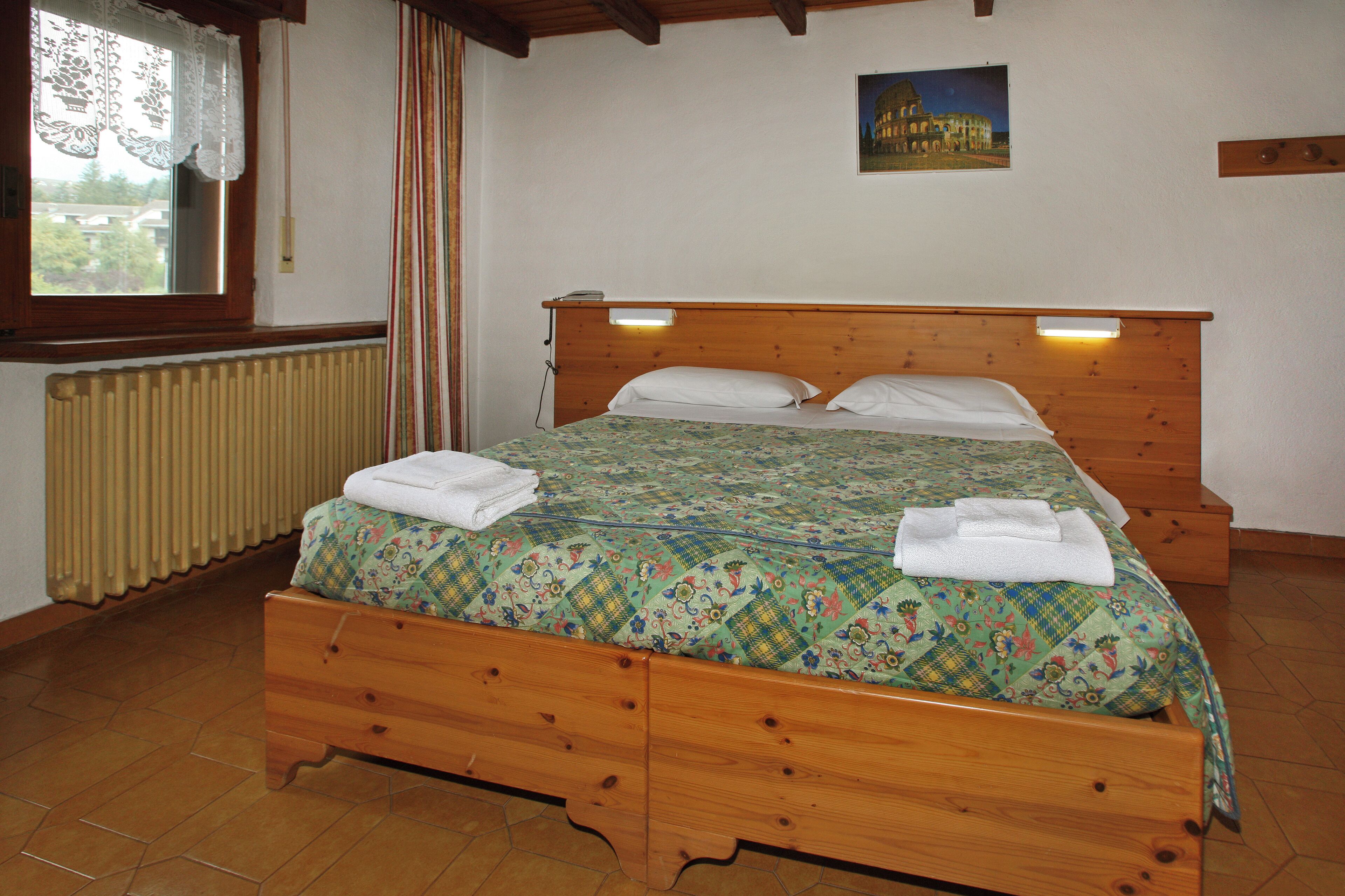 Un lit en bois avec une couvre-lit à motifs et deux oreillers, un radiateur en dessous, une fenêtre avec des rideaux et un tableau accroché au mur.