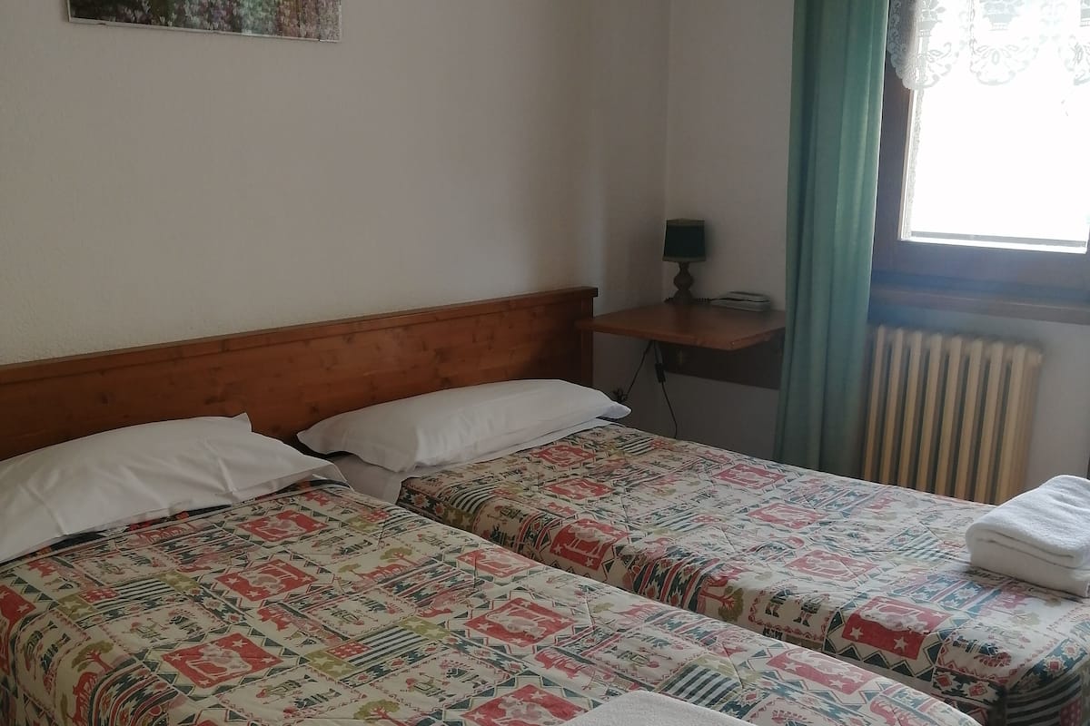 A room with two beds, a wooden headboard, a small table with a lamp, a radiator, and a window with curtains.