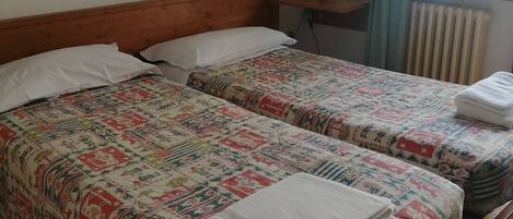 A room with two beds, a wooden headboard, a small table with a lamp, a radiator, and a window with curtains.