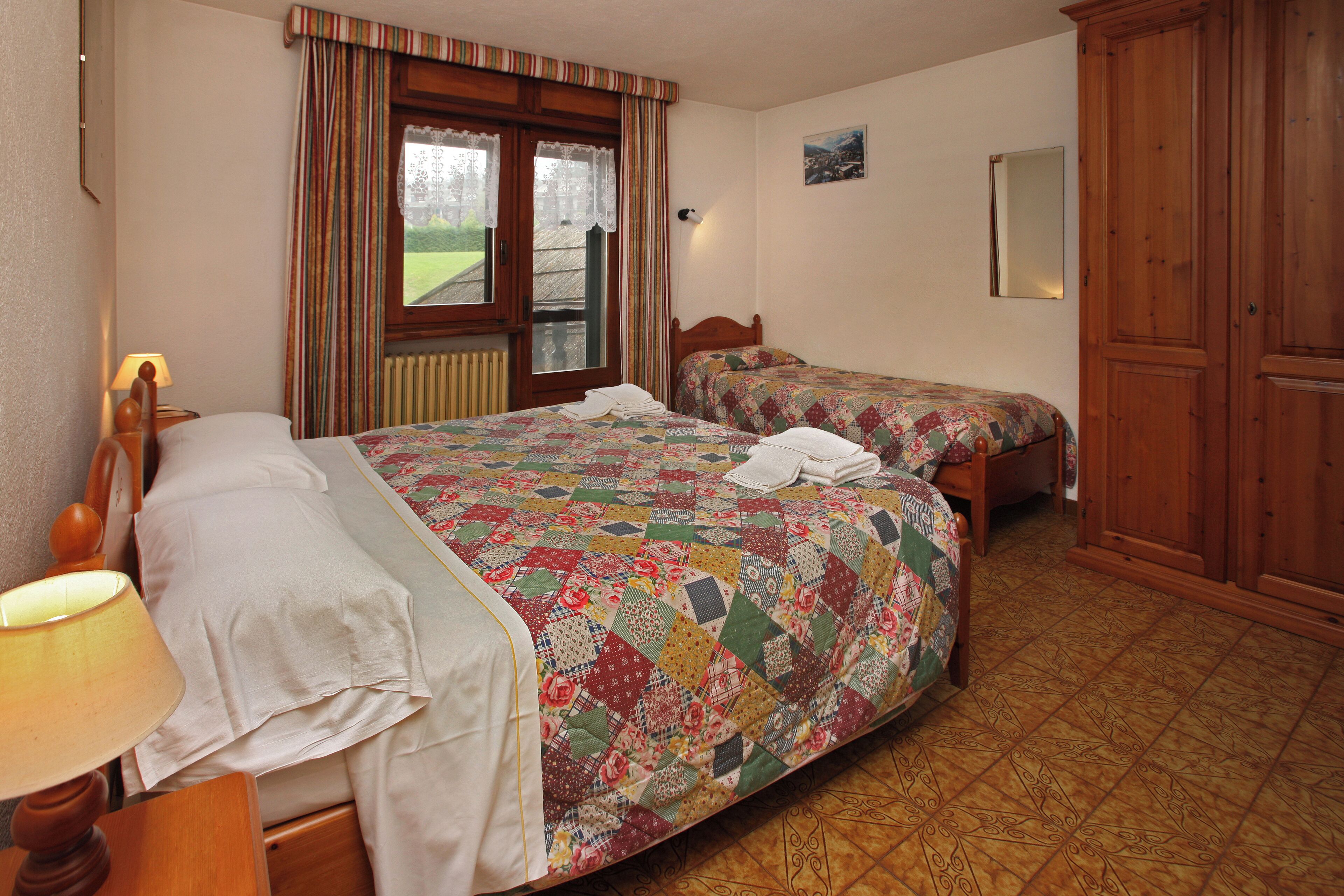 A bedroom with two beds, a wooden wardrobe, a window with curtains, and a lamp.