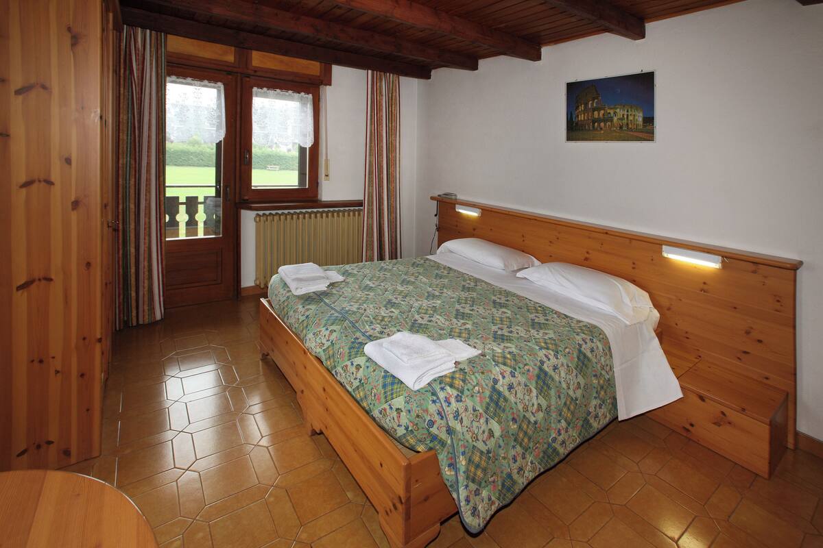 A wooden bed with a floral bedspread and white pillows, a wooden headboard, a window with curtains, and a painting of a building on the wall.