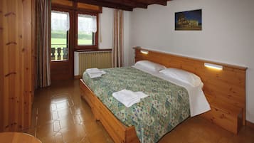 A wooden bed with a floral bedspread and white pillows, a wooden headboard, a window with curtains, and a painting of a building on the wall.