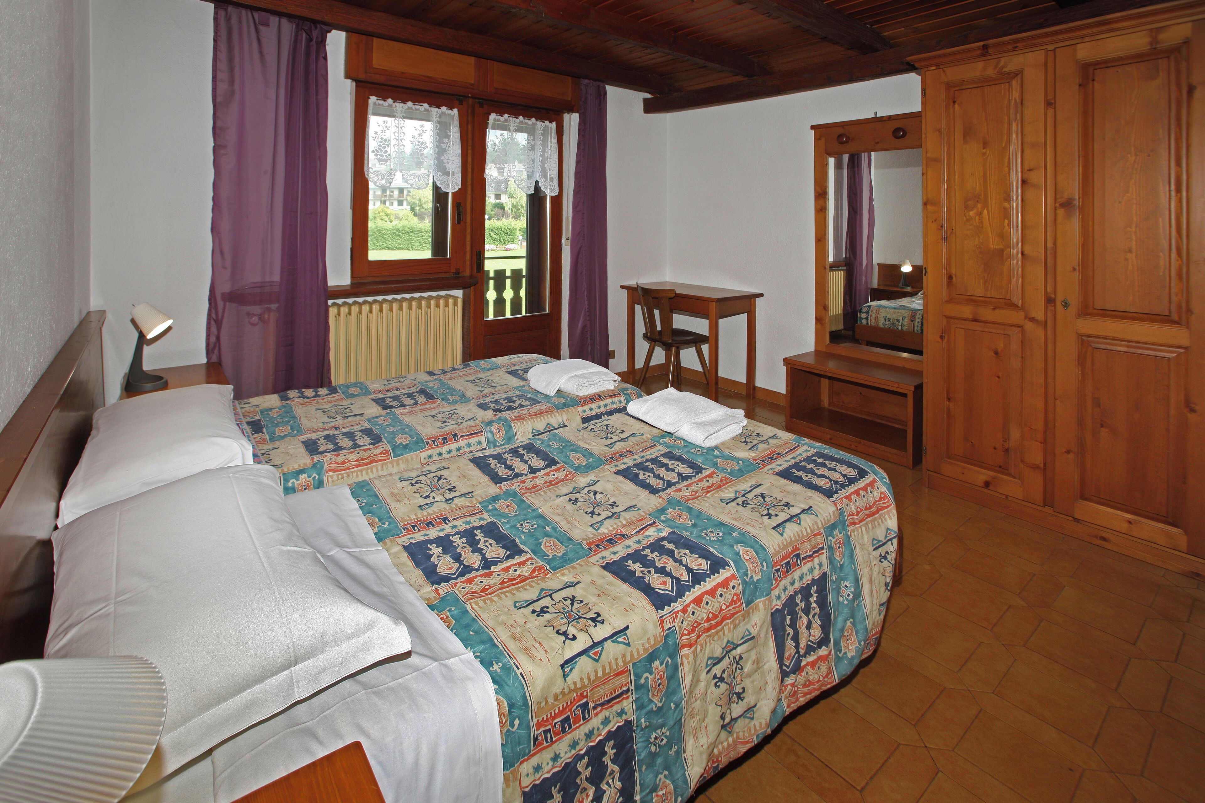 Un dormitorio con una cama grande, un armario de madera, una mesita pequeña y una ventana con cortinas.