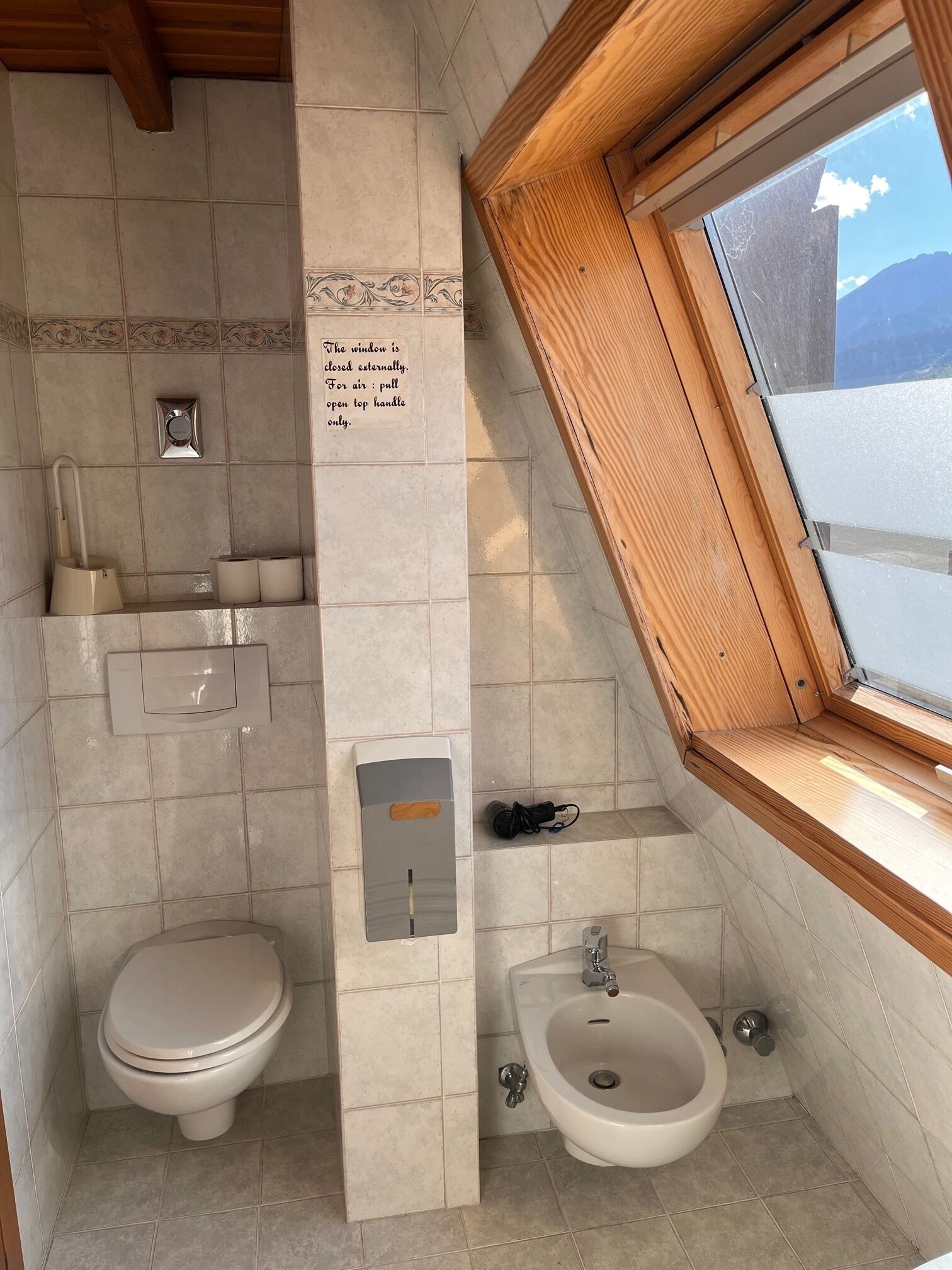 A bathroom with a toilet, bidet, and sink, a window with a wooden frame, and a wall-mounted air freshener.