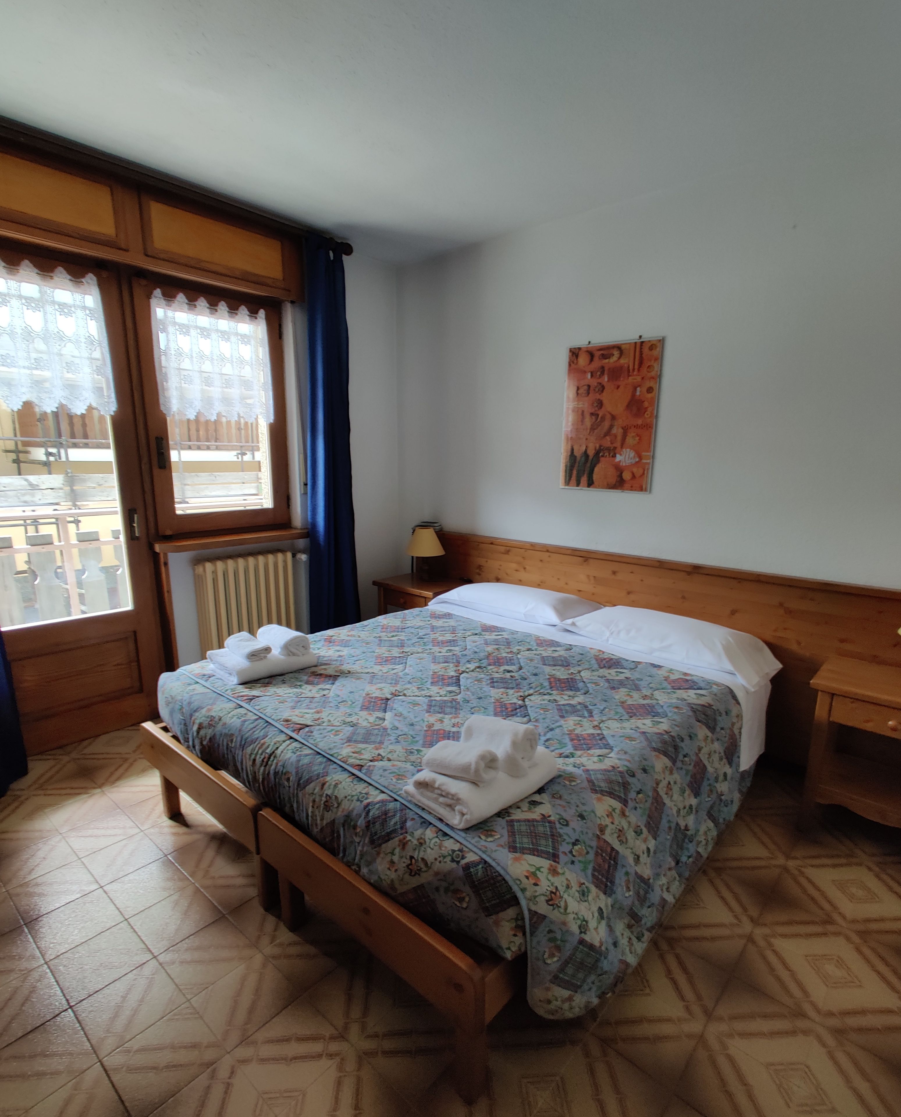 A hotel room with a wooden bed, a window with lace curtains, a radiator, a nightstand with a lamp, and a wall with a poster.