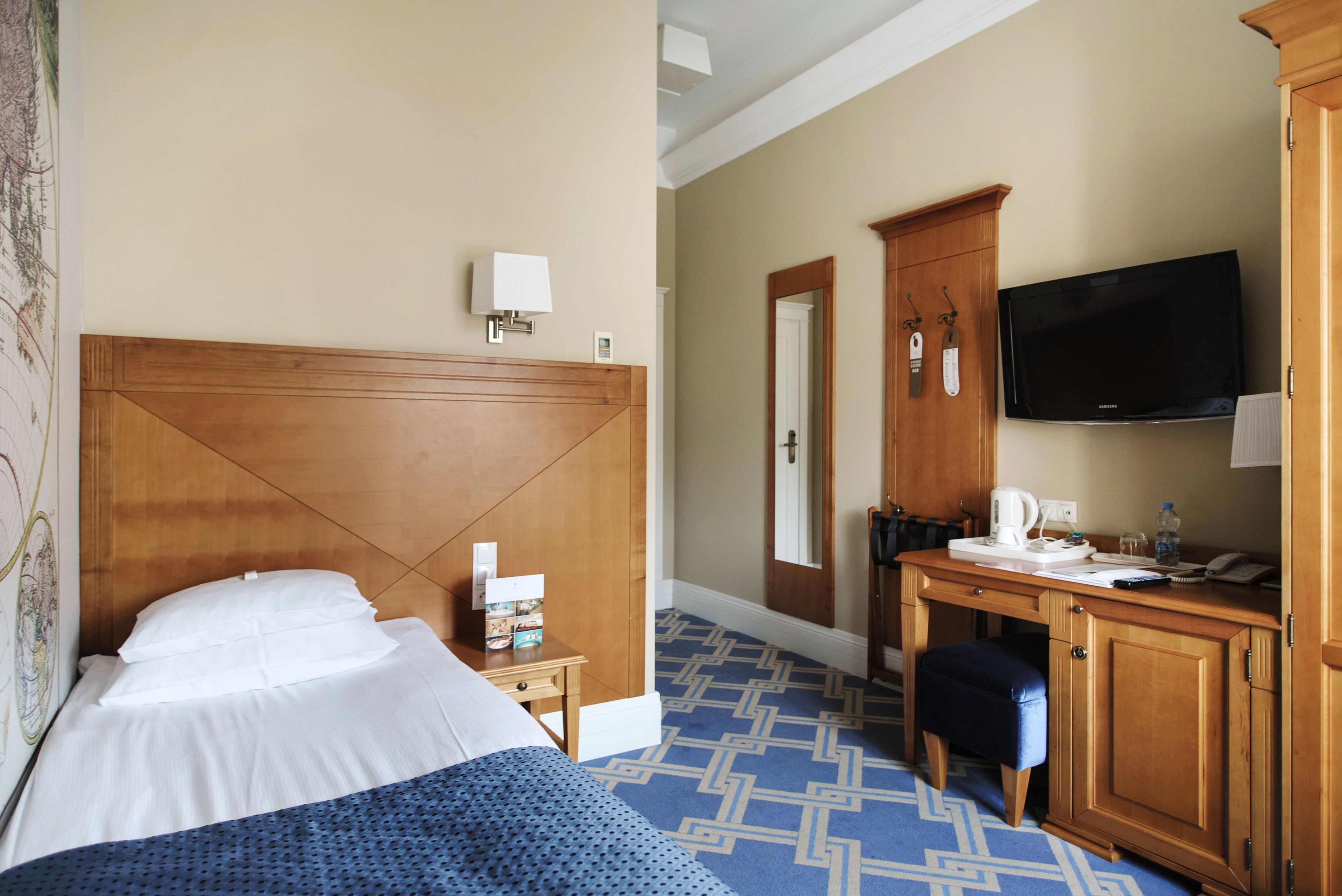 A hotel room with a wooden headboard, a bed with white linens, a desk with a television, and a patterned carpet.