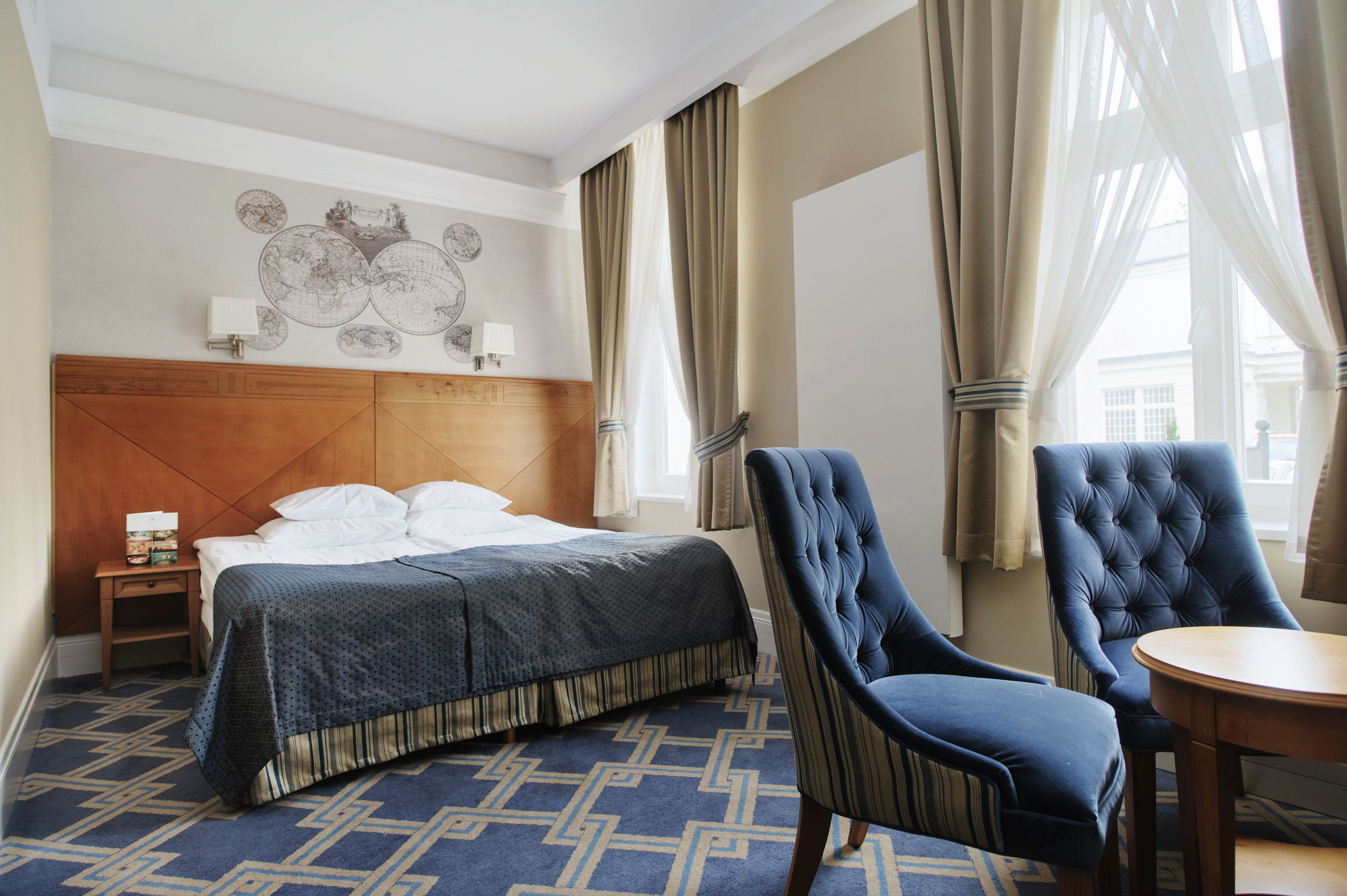 A hotel room with a large bed, a wooden headboard, a world map artwork, a blue upholstered chair, a round wooden table, and a patterned carpet.