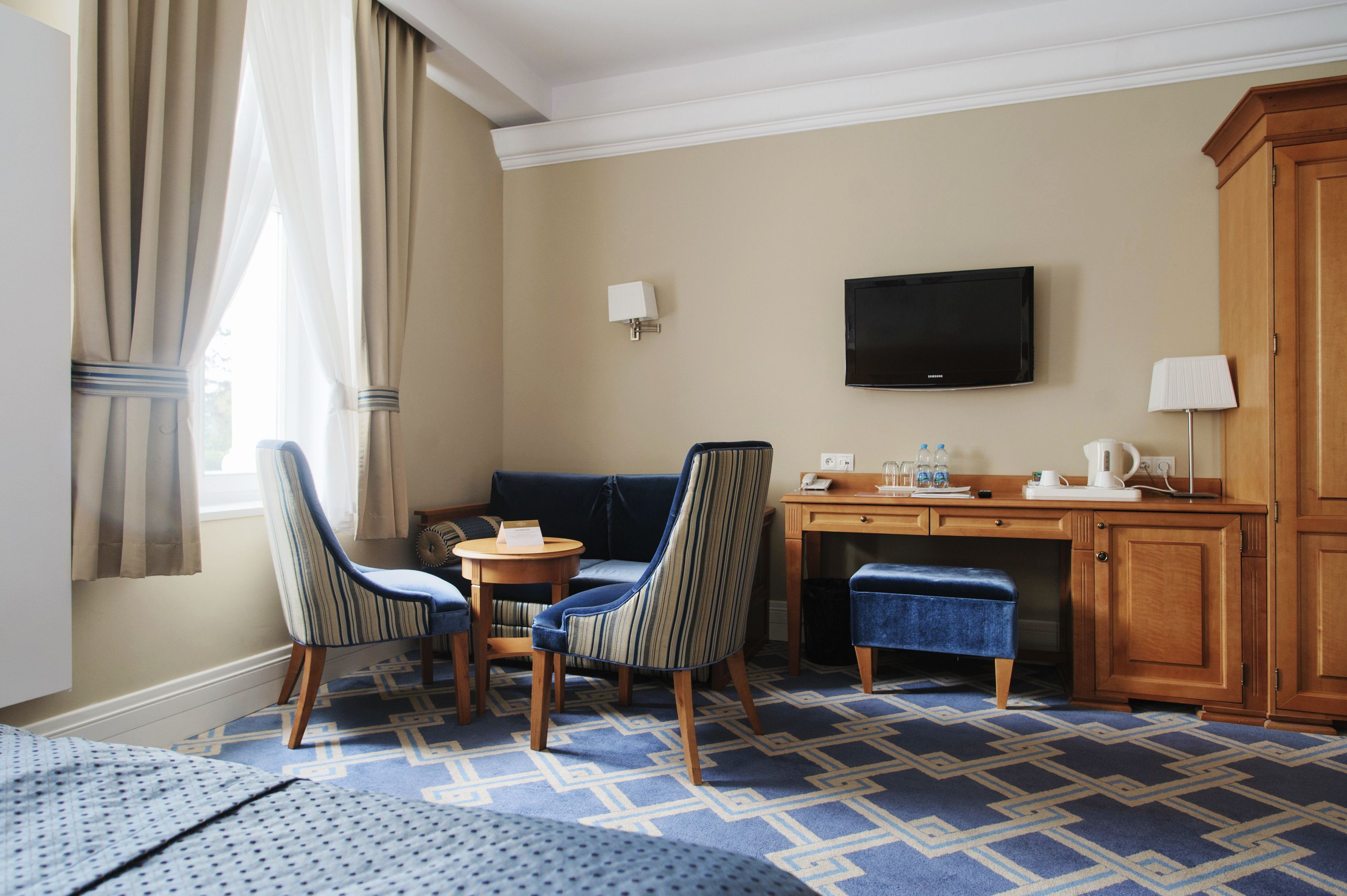 Een hotelkamer met een bank, twee gestreepte stoelen, een klein rond tafeltje, een bureau met een flatscreen televisie, een kast en een kamerbreed tapijt met dessin.