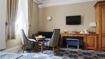 Een hotelkamer met een bank, twee gestreepte stoelen, een klein rond tafeltje, een bureau met een flatscreen televisie, een kast en een kamerbreed tapijt met dessin.