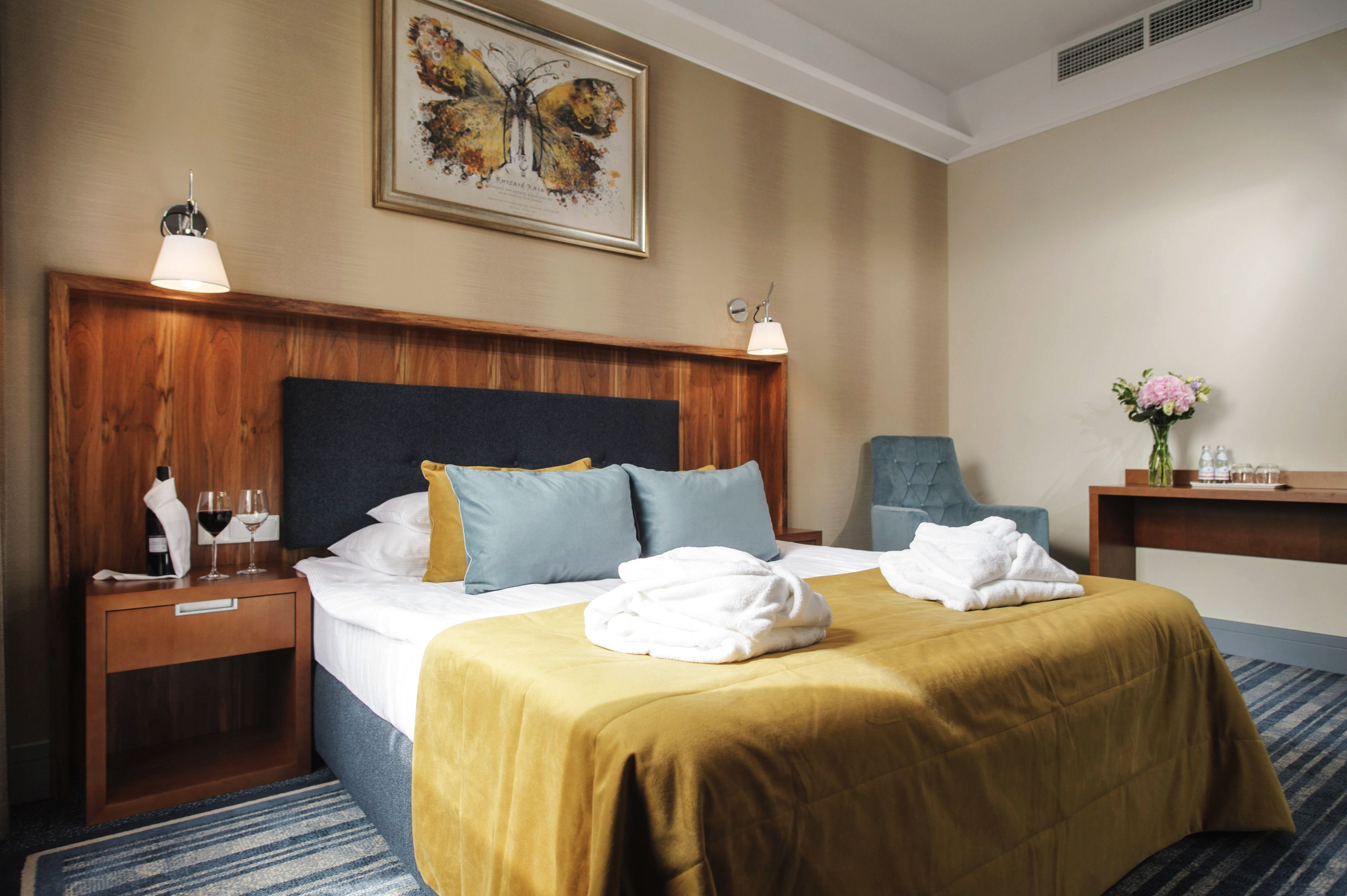 Habitación de hotel con una cama grande, cabecera de madera, dos mesitas de noche, una mesita auxiliar, una obra de arte con mariposas en marco, un jarrón con flores y una silla azul.