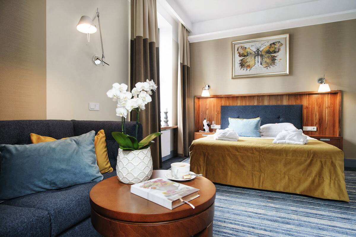 A hotel room with a bed, a sofa, a coffee table with a book and flowers, and a painting on the wall.