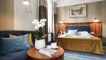 Quarto de hotel com cama, sofá, mesa de centro com livro e flores, e um quadro na parede.