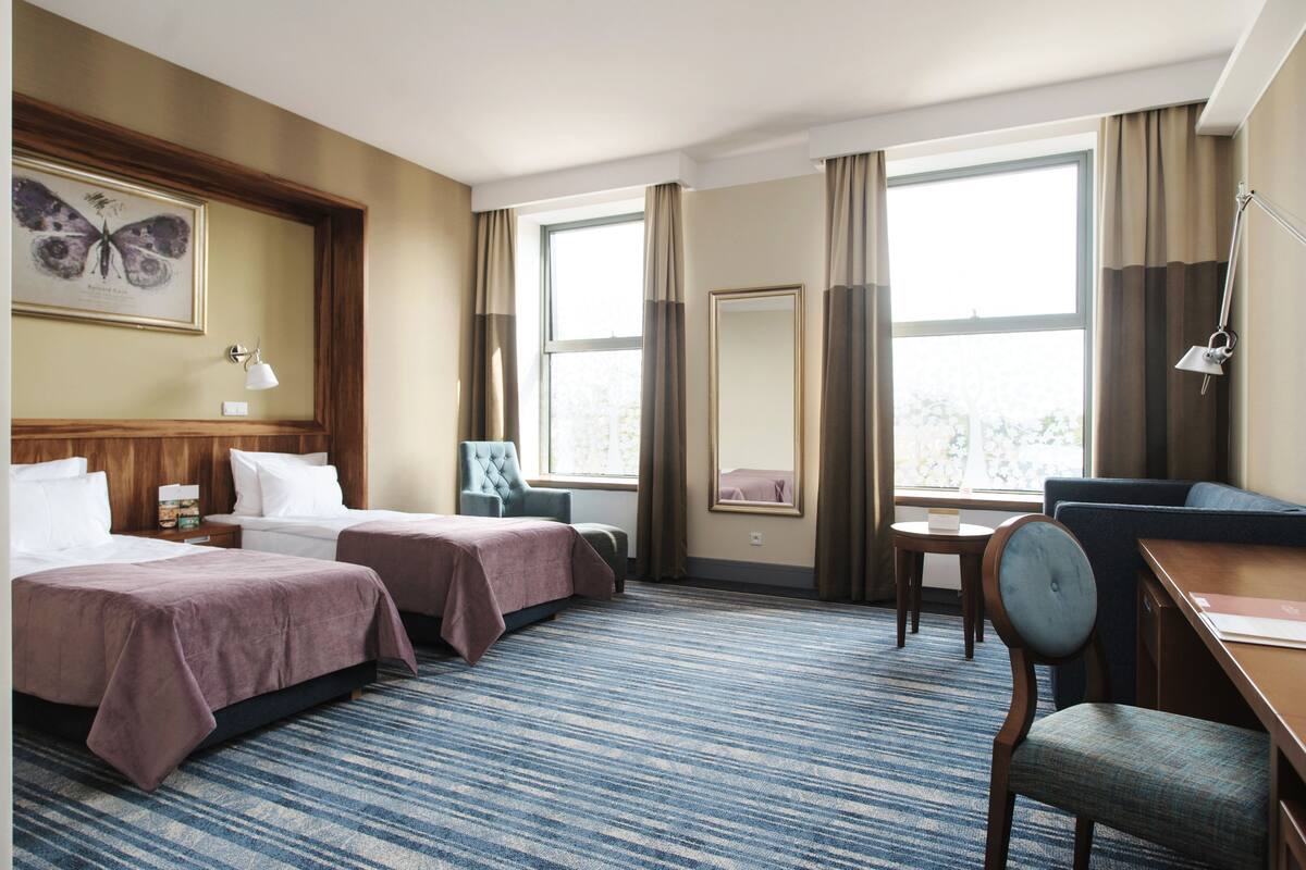 A hotel room with two beds, a desk, a chair, a window with curtains, a framed butterfly artwork, and a lamp.