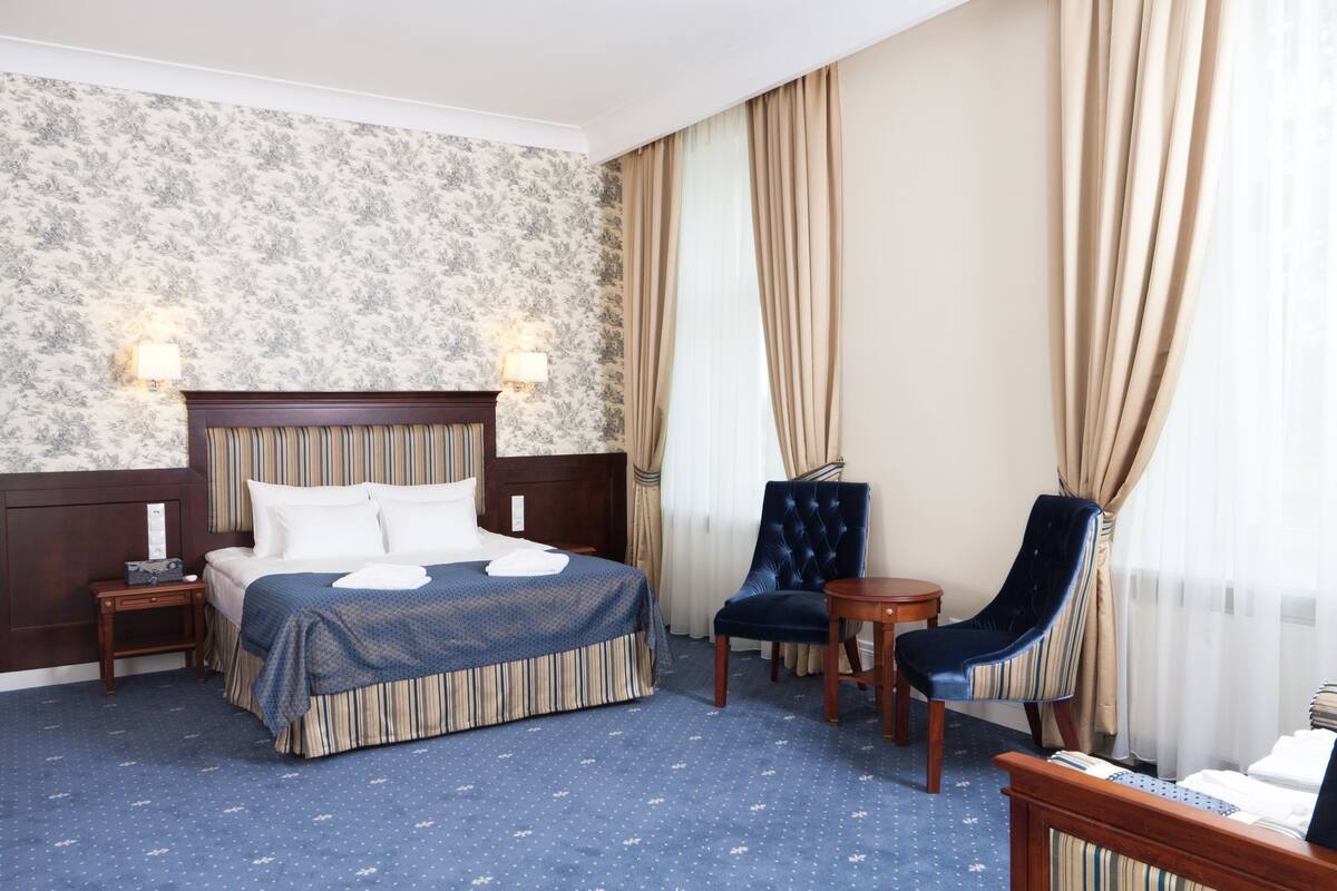A hotel room with a large bed, two bedside tables, a chair, a small table, and a window with curtains.