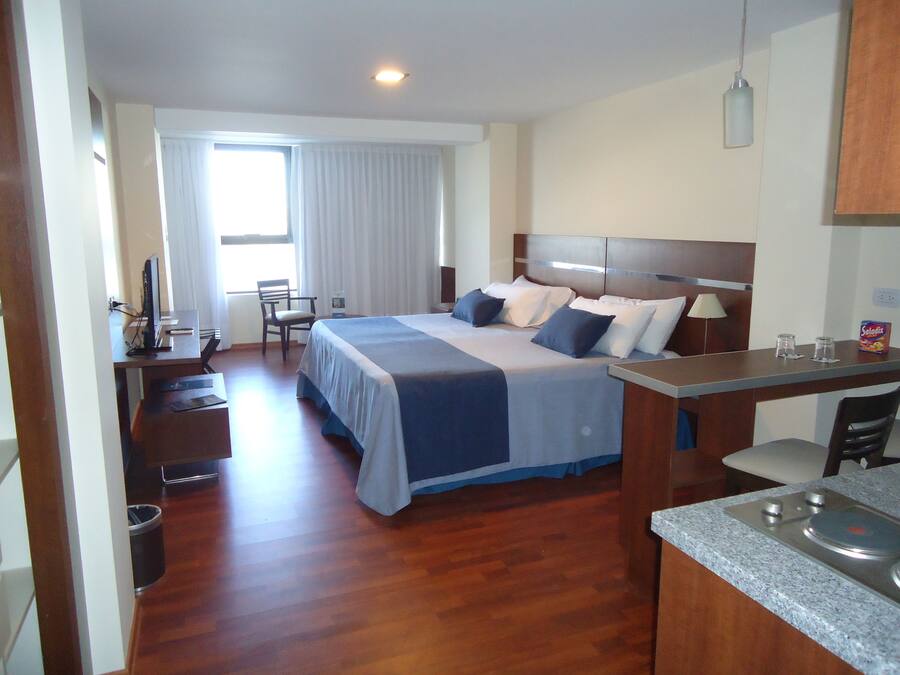 A hotel room with a bed, a desk with a computer, a chair, a window with curtains, and a kitchenette.