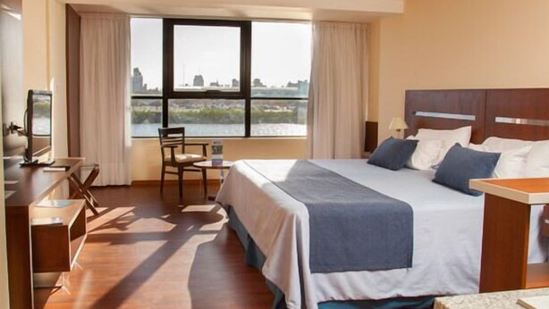 Habitación de hotel con una cama grande, un escritorio con una silla, un televisor y una ventana con vista a la ciudad.