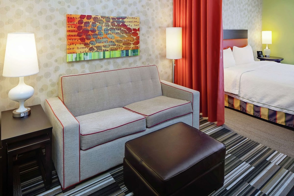 A hotel room with a sofa, a small table, a bed, and a wall with a colorful artwork.