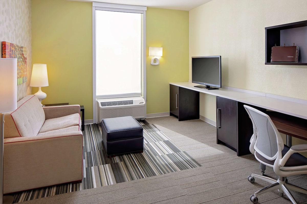 A hotel room with a sofa, a desk with a computer, a TV, and a window with blinds.