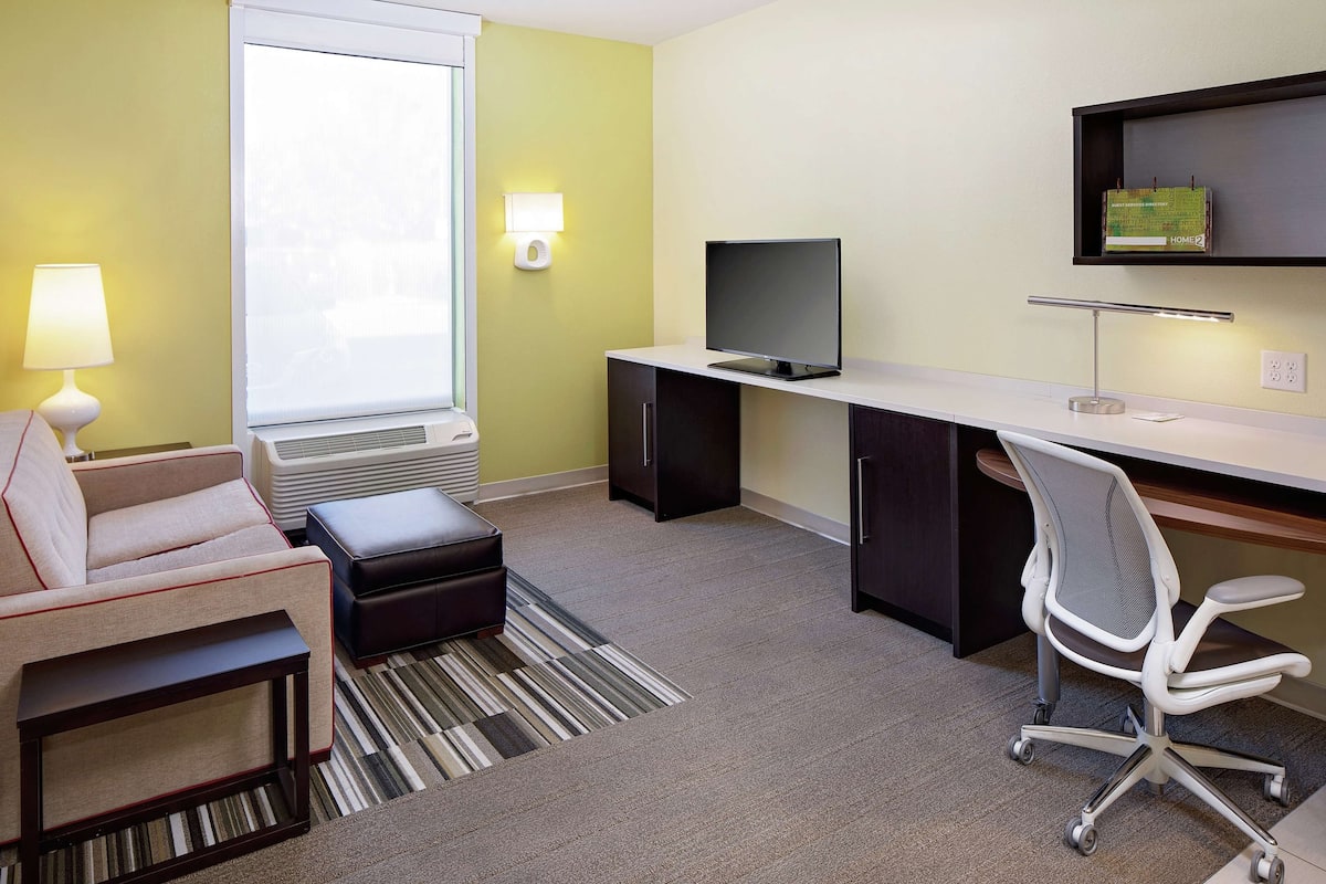 A modern hotel room with a sofa, a desk with a computer, a TV, and a lamp.