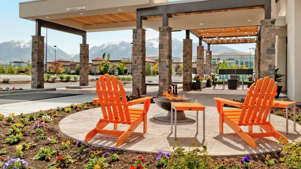 An outdoor seating area with two orange Adirondack chairs and a fire pit, surrounded by a landscaped garden with colorful flowers. - Home2 Suites by Hilton Salt Lake City/South Jordan, UT (South Jordan)