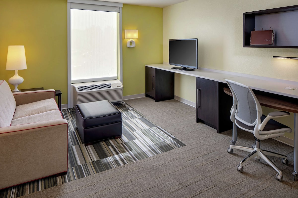 A hotel room with a sofa, a desk, a chair, a TV, and a window with blinds.