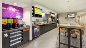 A modern hotel kitchen with a coffee station, a trash bin, and a long dining table with chairs. - Home2 Suites by Hilton Salt Lake City/South Jordan, UT (South Jordan)