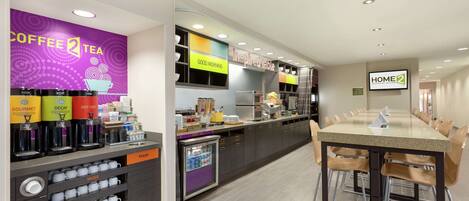 A modern hotel kitchen with a coffee station, a trash bin, and a long dining table with chairs.