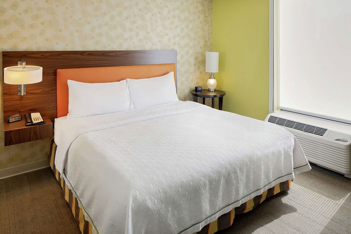 A hotel room with a bed, bedside table with a lamp, a phone, a small table with a clock, and a window with an air conditioner.