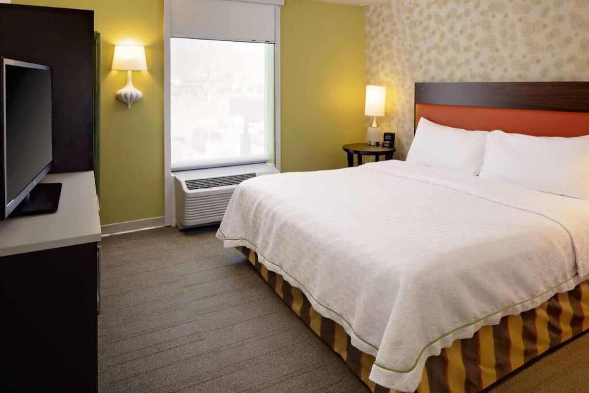 A hotel room with a bed, a television, a window with blinds, a wall-mounted lamp, and a small table with a lamp.