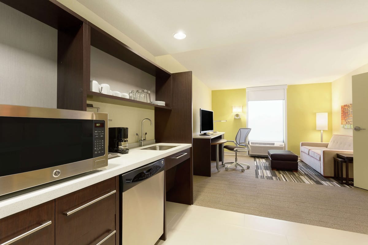 A modern kitchen with a built-in microwave, dishwasher, and a coffee maker. There is a dining area with a table and chairs, and a living area with a sofa and a TV.