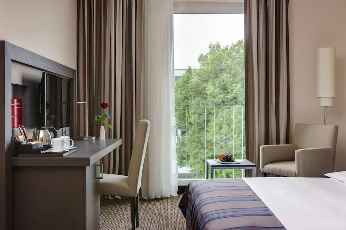 A hotel room with a bed, a desk with a TV, a chair, a small table with fruit, and a balcony with a railing.