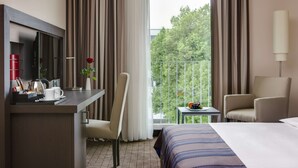 A hotel room with a bed, a desk with a computer, a chair, a small table with fruit, and a balcony with a view of trees. - IntercityHotel Ingolstadt (Ingolstadt)