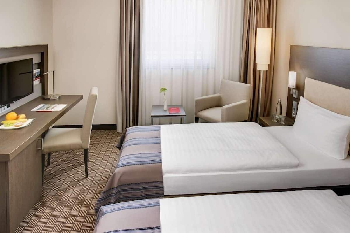 A hotel room with a bed, a desk with a TV, a chair, and a small table with a plant.