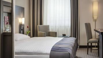 Une chambre d’hôtel avec un grand lit, un bureau, une chaise, une lampe et une vue sur l’extérieur à travers des rideaux légers.