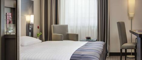 Habitación de hotel con una cama grande, un escritorio, una silla, una lámpara y vistas al exterior a través de cortinas translúcidas.