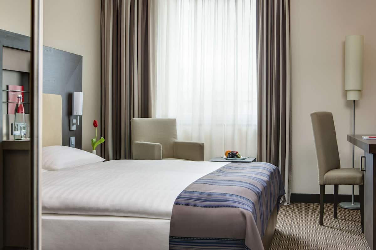 A hotel room with a large bed, a desk, a chair, and a window with curtains.