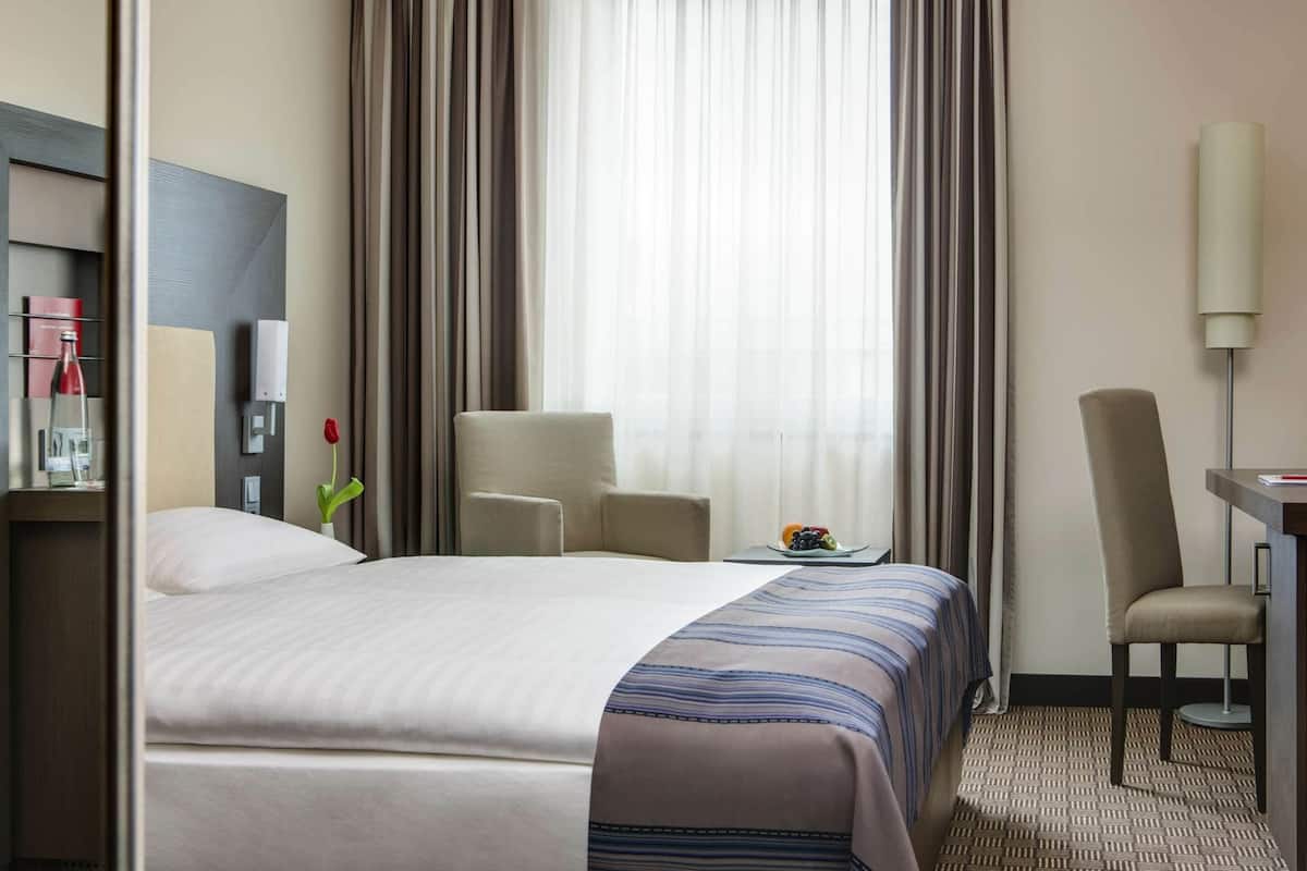 A hotel room with a bed, two chairs, a desk with a lamp, a small table with fruit, and a window with curtains.