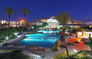 A large outdoor swimming pool with multiple water features, surrounded by palm trees and lounge chairs, with a building in the background. - Joya Paradise (Djerba Midun)