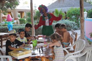 Eine Gruppe Kinder sitzt an einem Tisch im Freien, während ein Maskottchen in der Nähe steht.